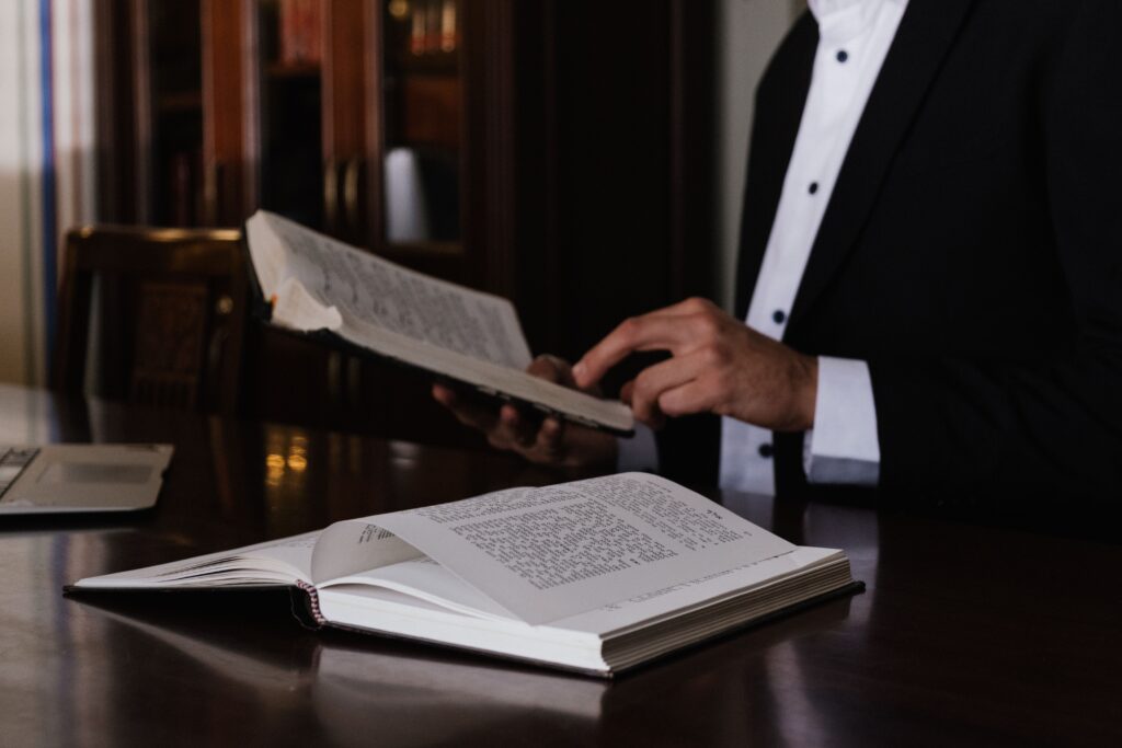A man in a suit reading religious books at a wooden table indoors.